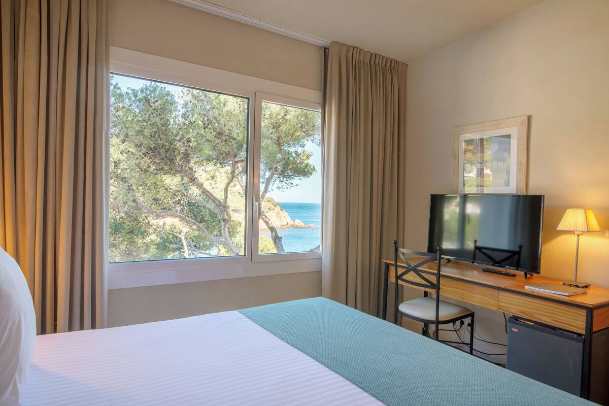 A hotel room with a bed, a desk with a TV, a chair, and a view of the sea through large windows.