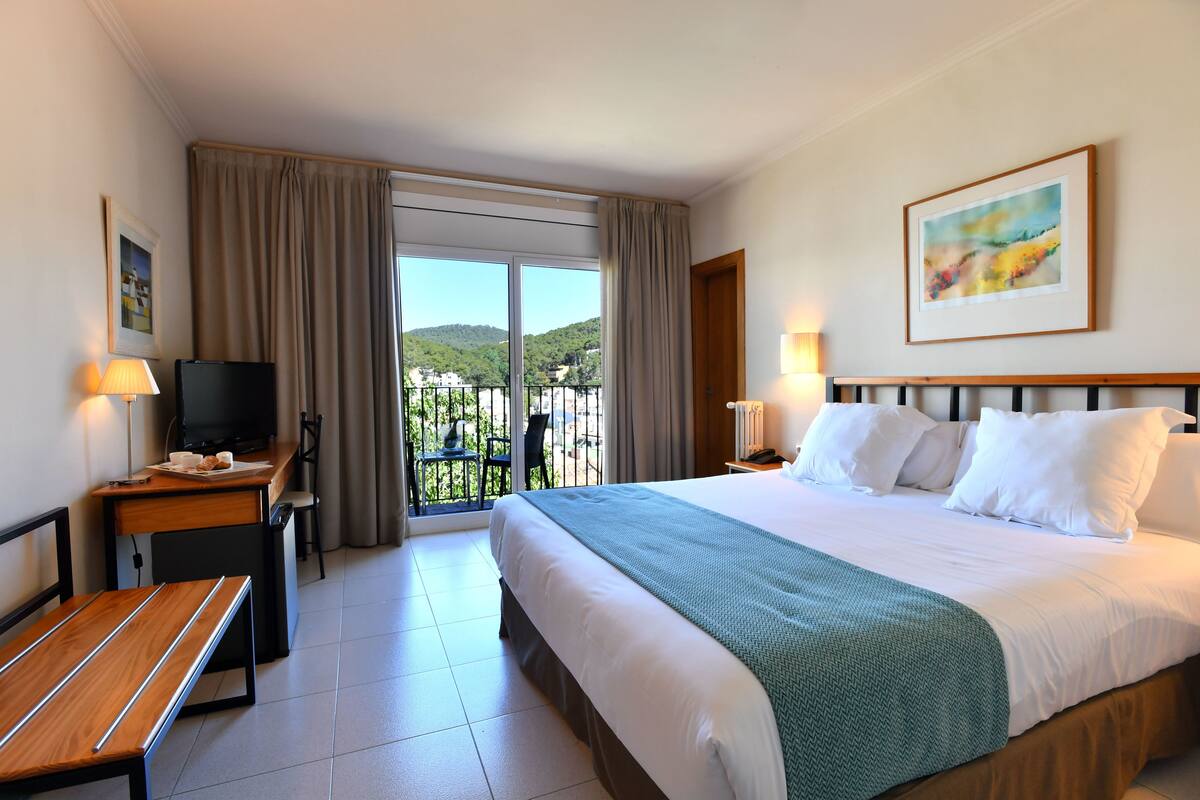A hotel room with a bed, a desk with a television, a chair, a bench, and a balcony with a view of greenery.