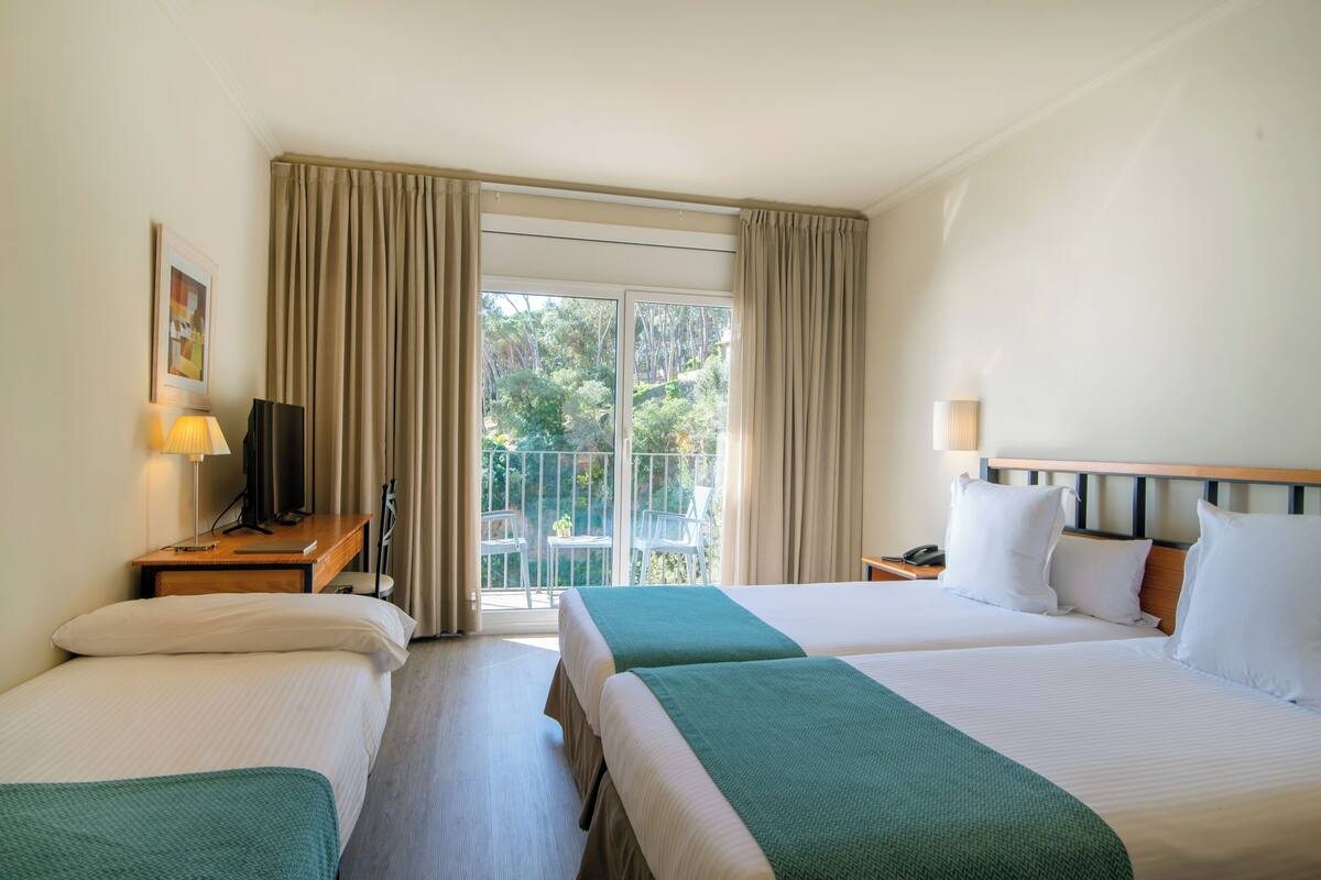 A hotel room with two beds, a desk with a lamp, a TV, and a balcony with chairs.