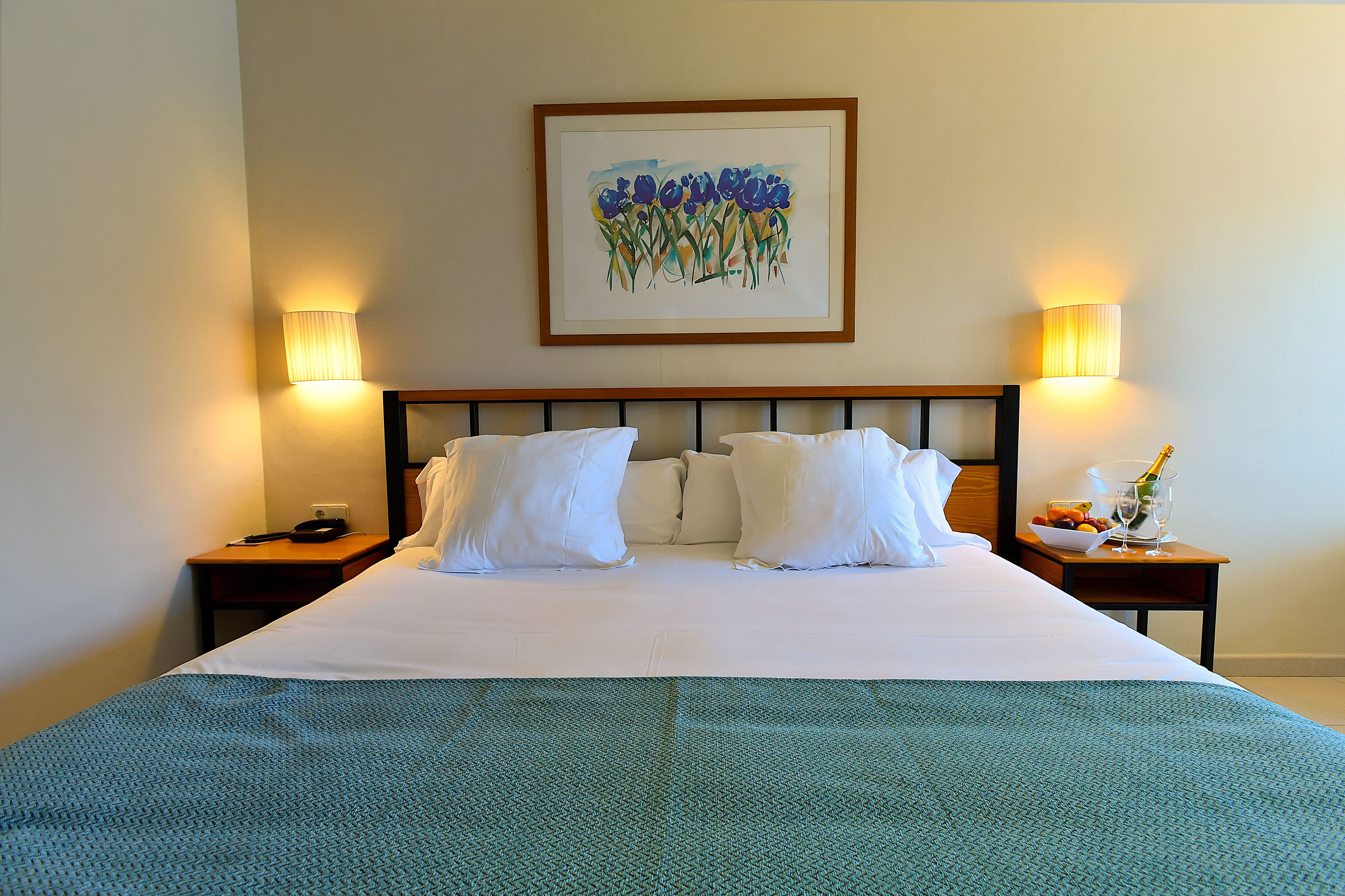 A neatly made bed with white linens and a green patterned bedspread, flanked by two bedside tables with lamps, a framed artwork above the bed, and a small table with a bottle and fruit.