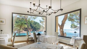 Un comedor con mesas dispuestas para dos, ventanas amplias con vistas al mar y árboles, y una lámpara moderna.