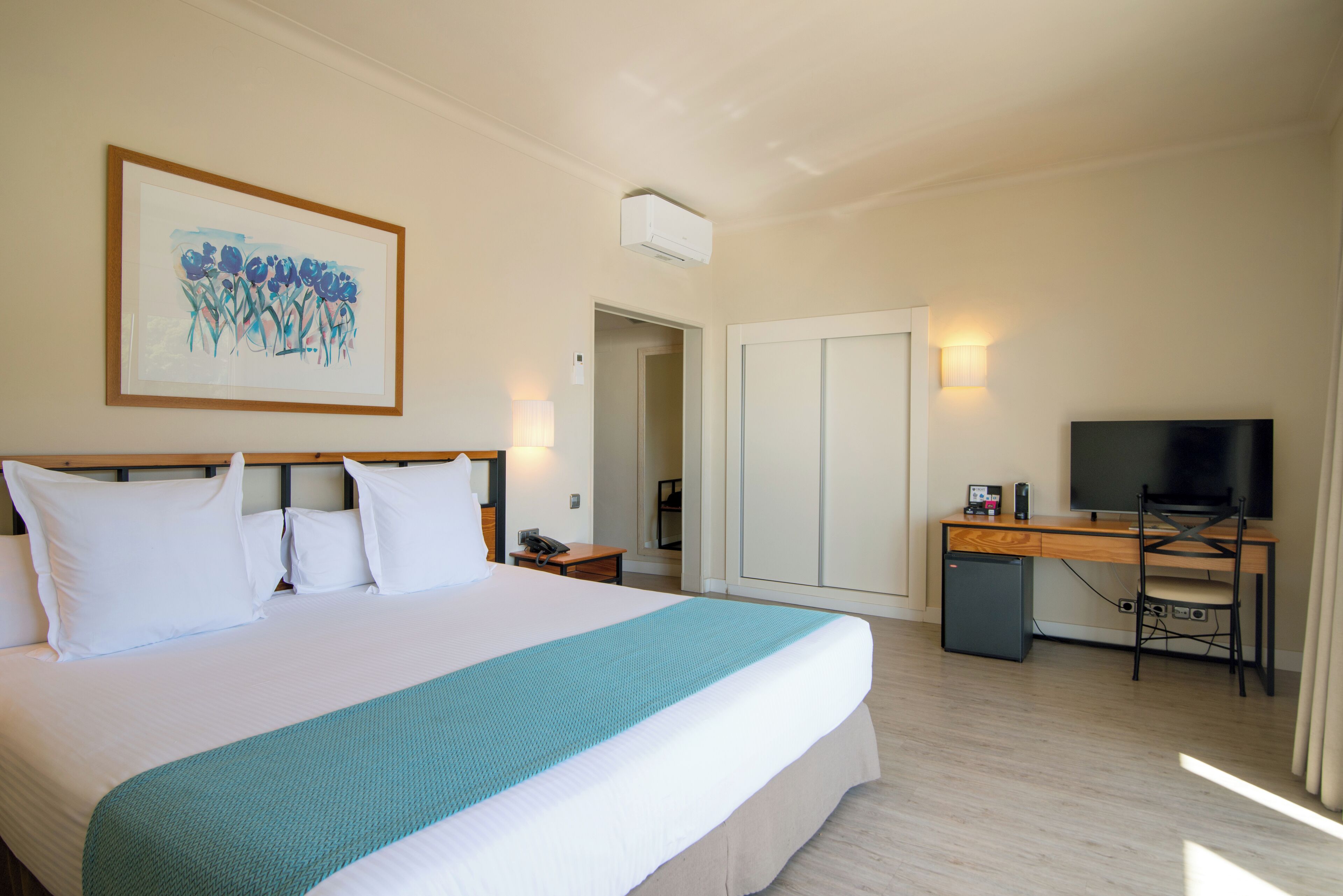 A hotel room with a large bed, a desk with a computer, a TV, and a framed artwork on the wall.