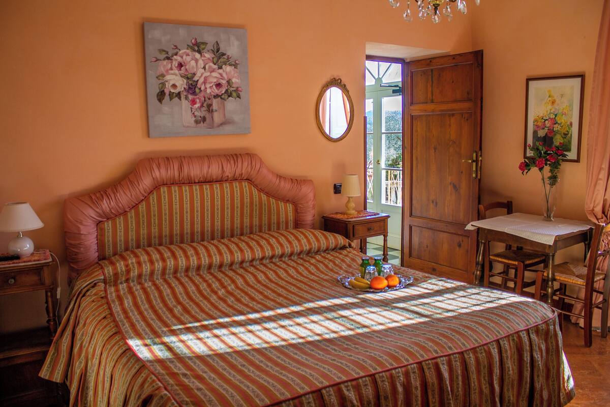 A bedroom with a large bed, a bedside table with a lamp, a small table with a vase of flowers, and a door leading to a balcony.