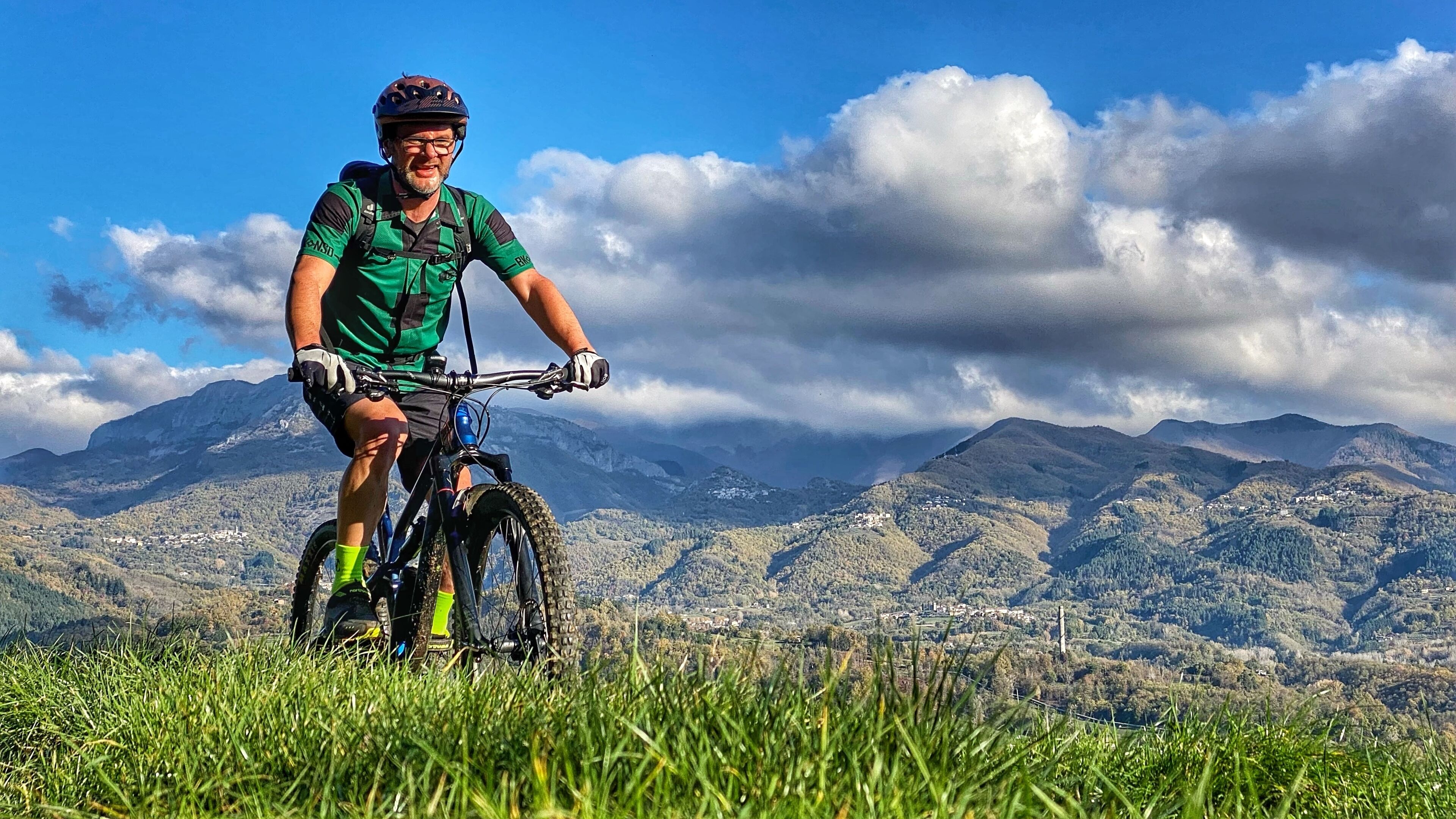 Uma pessoa andando de bicicleta de montanha em uma colina gramada, com uma paisagem montanhosa ao fundo.