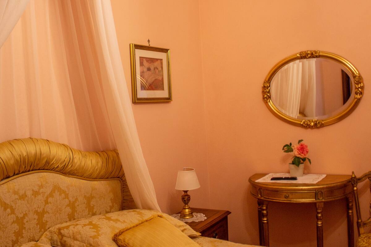 A bedroom with a canopy bed, a bedside table with a lamp, a mirror, and a small table with a vase.