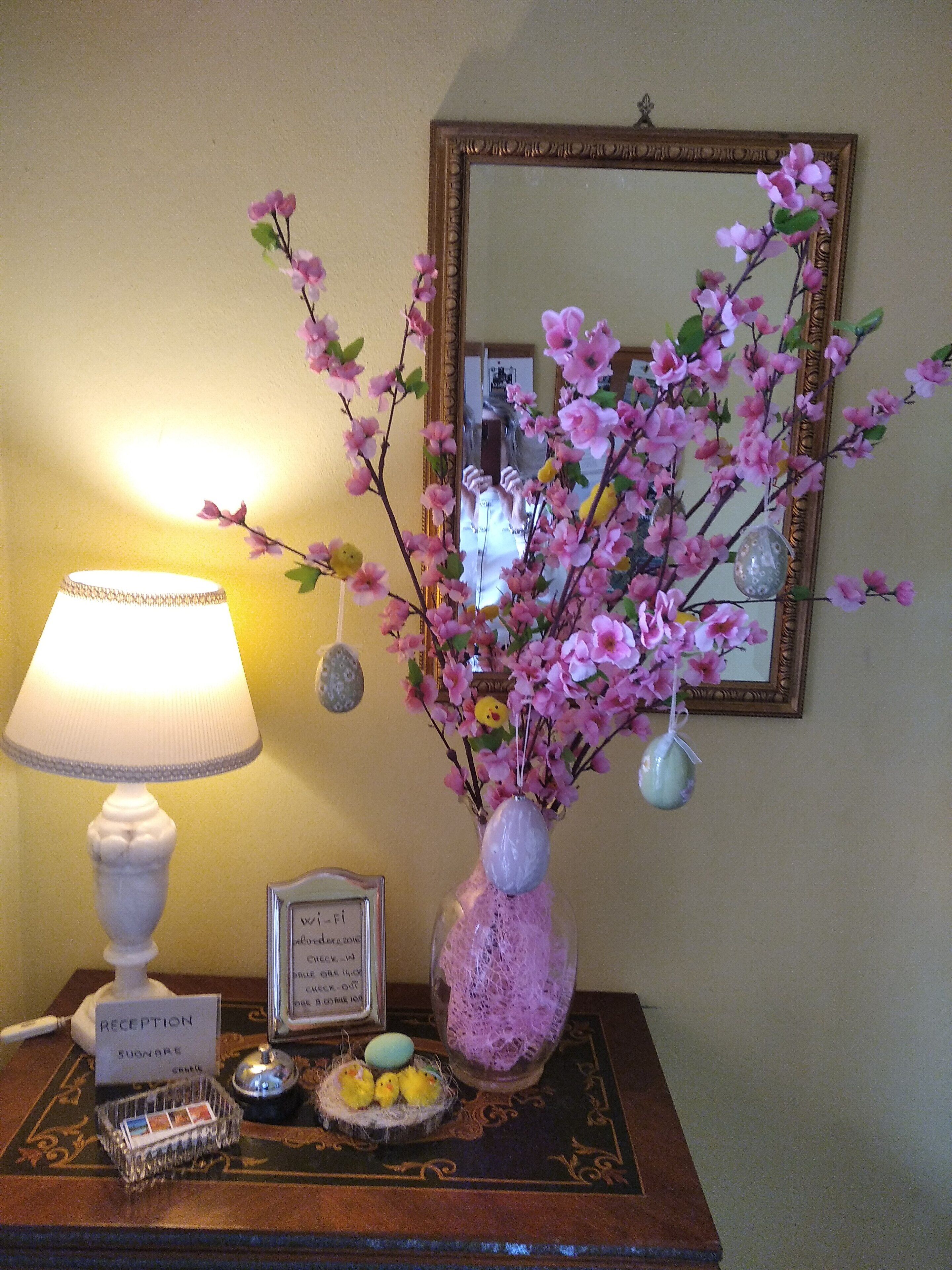 Ein Empfangsschalter mit einer Lampe, einer Vase mit rosa Blumen, einem gerahmten Schild und einem dekorativen Gegenstand mit Hänschen-Figuren.