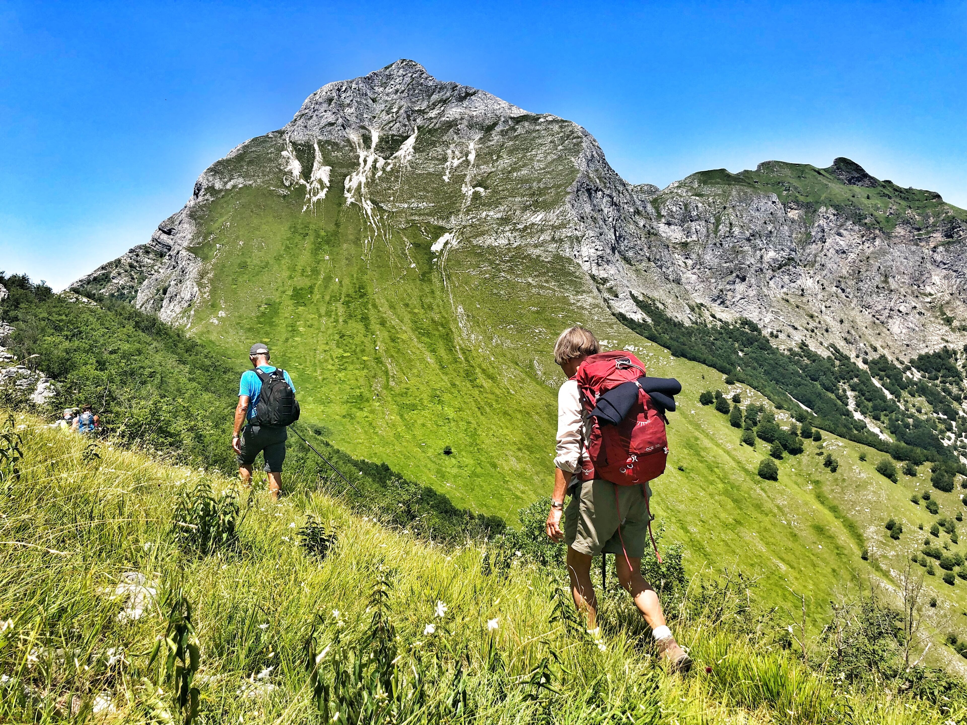 Two hikers on a grassy mountain trail with rocky outcrops and a prominent mountain in the background.