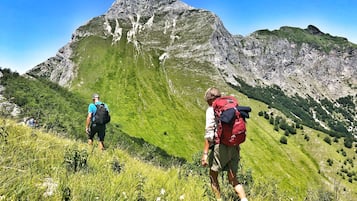 Due escursionisti su un sentiero montano erboso, con affioramenti rocciosi e una montagna imponente sullo sfondo.
