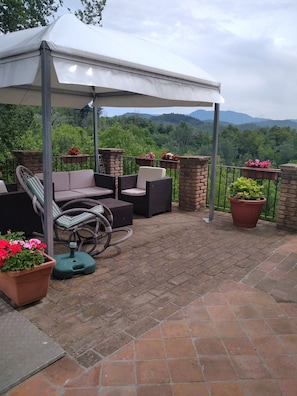 A covered patio area with outdoor furniture, potted plants, and a view of a mountainous landscape.