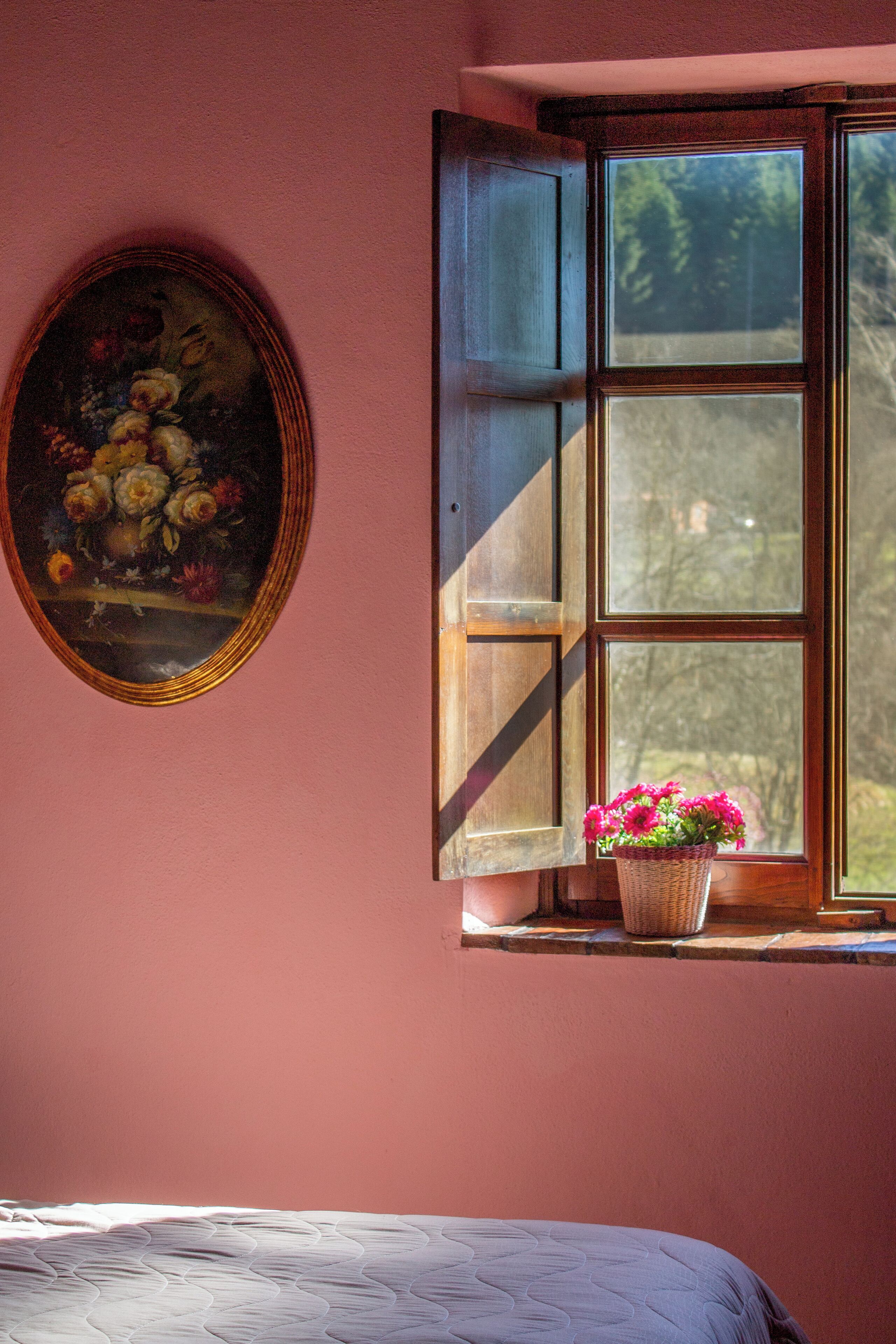 Una habitación con cama, una ventana con contraventanas abiertas y un cuadro en la pared.