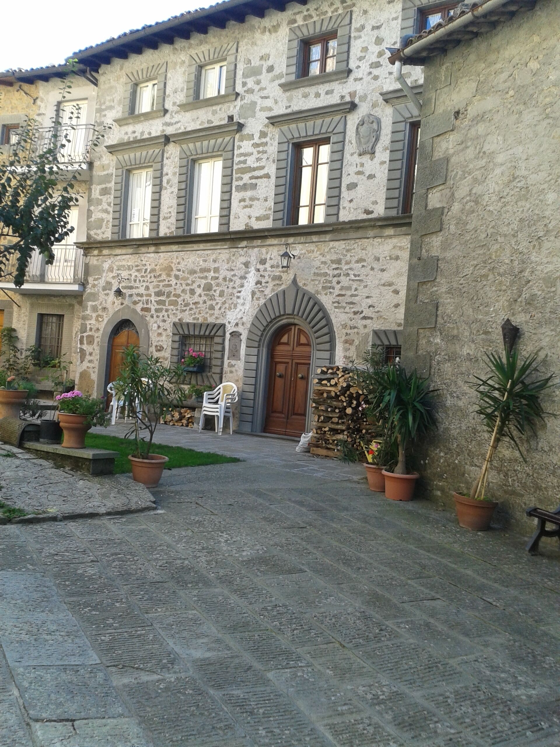Un edificio de piedra con una entrada central arqueada, plantas en macetas y un pequeño espacio con mesas al aire libre.