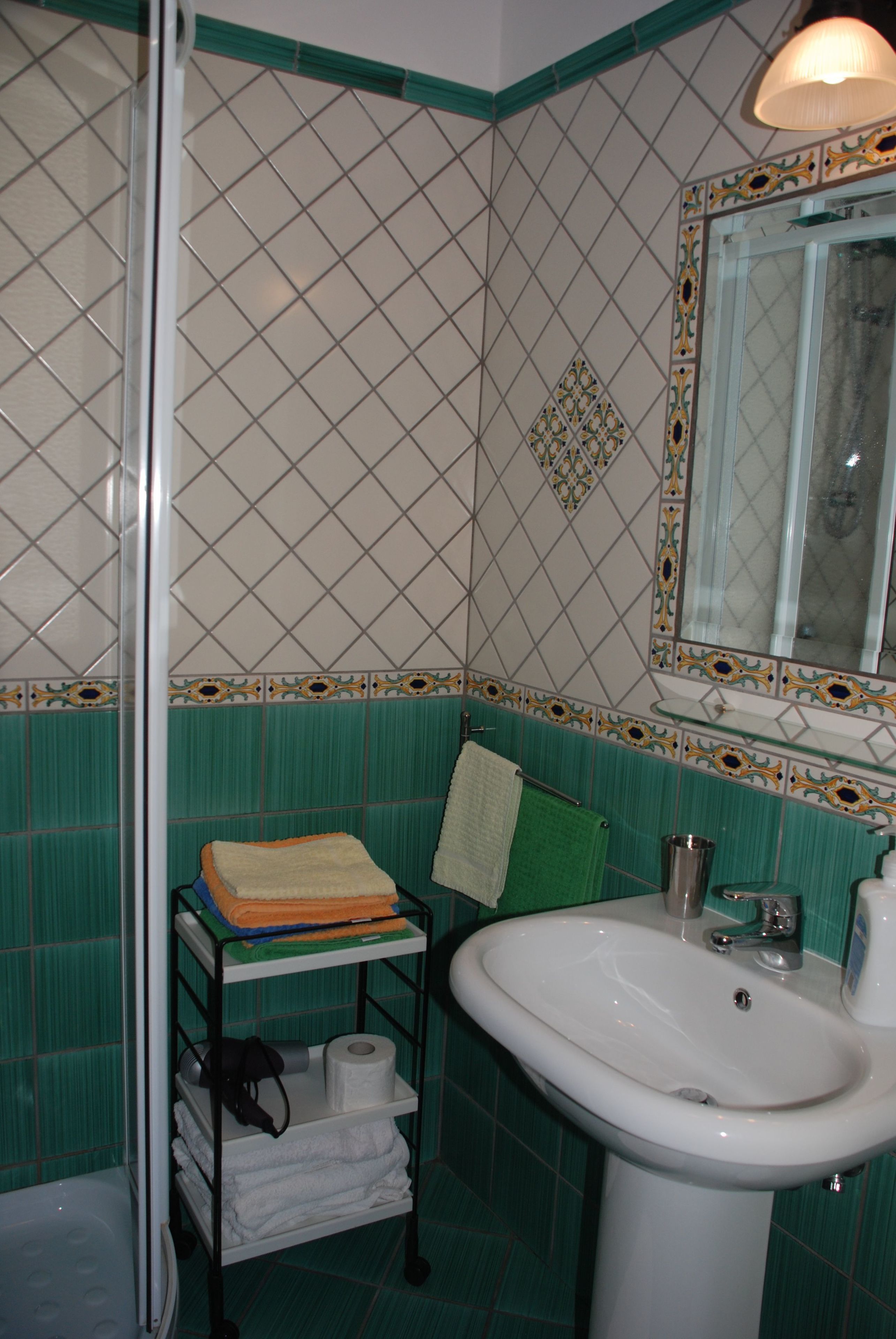 A bathroom with a white sink, green tiled walls, a glass shower enclosure, and a towel rack with folded towels.