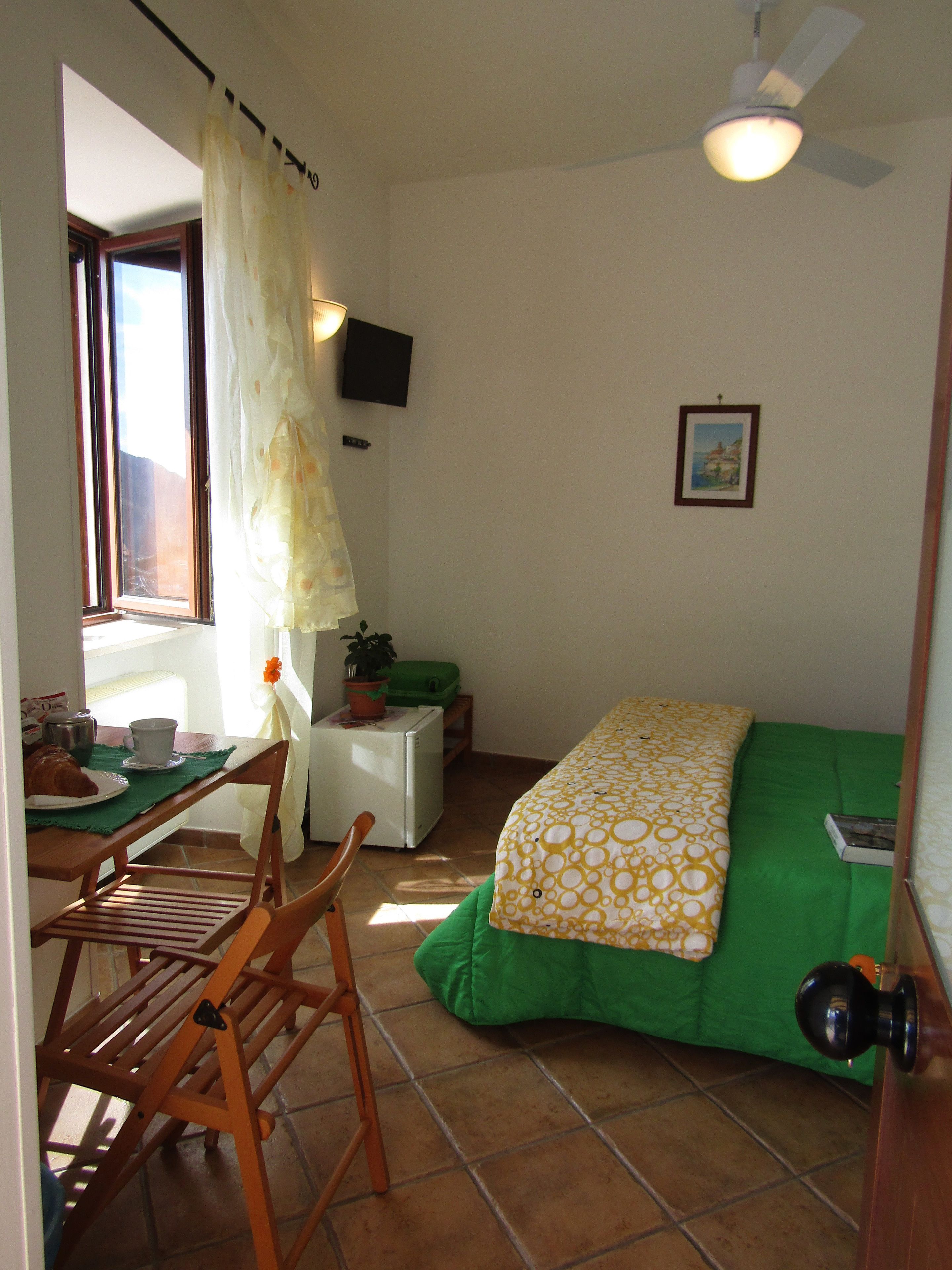 Una habitación pequeña y acogedora con cama, una mesa con silla, ventana con cortinas, ventilador de techo y televisor instalado en la pared.