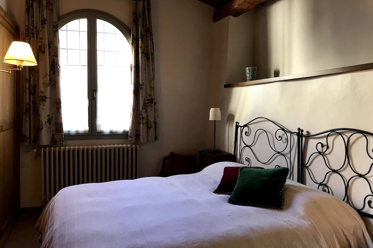 Un dormitorio con una cama grande, una ventana con cortinas, un radiador, una mesita de noche con lámpara y un estante sobre la cama.