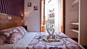 Una habitación de hotel con una cama, cabecera de madera, ventana con vistas a edificios y un armario blanco.