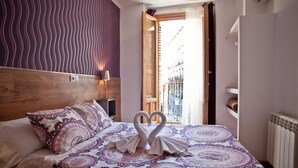 Habitación de hotel con cama, cabecera de madera, mesita de noche, lámpara y vista a la calle a través de una puerta abierta.