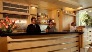 A reception desk with a wooden finish, a plant on the left, and a window with a view of greenery. - Hotel Alka Classic (New Delhi)