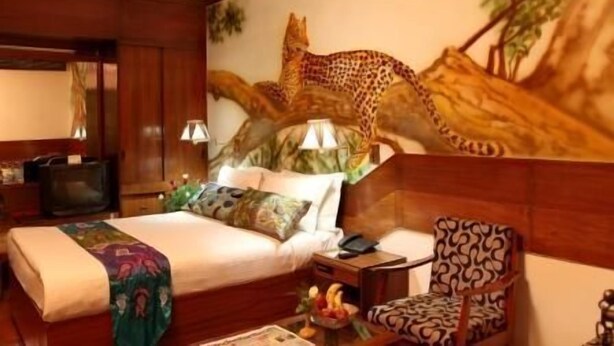 A hotel room with a large leopard mural on the wall, a bed with a patterned bedspread, a wooden nightstand, a chair, and a small table with fruit. - Hotel Alka Classic (New Delhi)