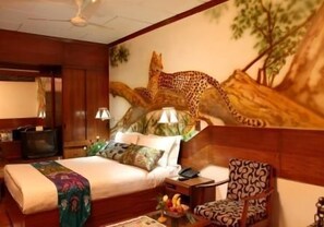 Quarto de hotel com um grande mural de leopardo na parede, uma cama com colcha estampada, uma mesinha de cabeceira de madeira, uma cadeira e uma pequena mesa com frutas.
