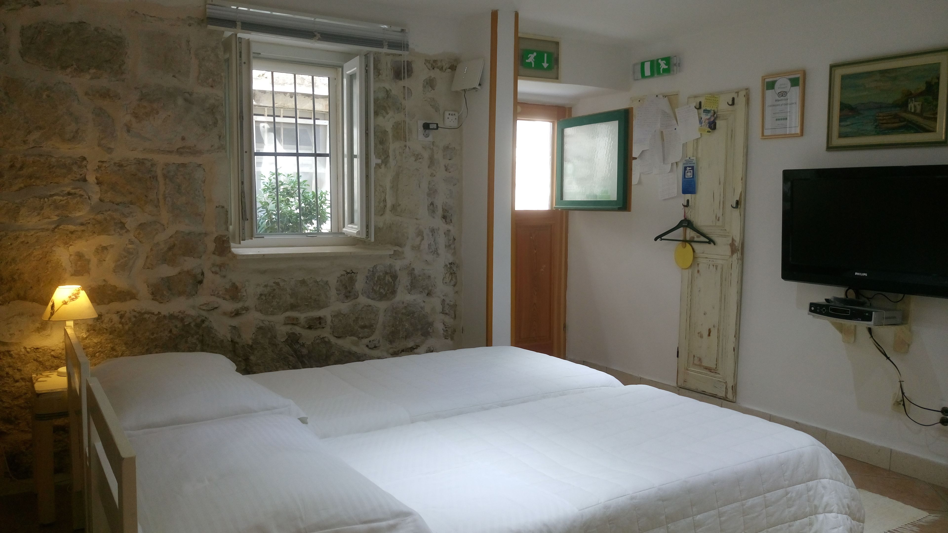 A room with a stone wall, a bed with white bedding, a window with bars, a door with a green exit sign, a wall-mounted TV, and a lamp.