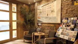 Une pièce chaleureuse aux murs en bois, avec une grande fenêtre, un coin salon composé de deux fauteuils, une petite table sur laquelle est posé un vase, et un présentoir mural où sont exposés des livres et des brochures.