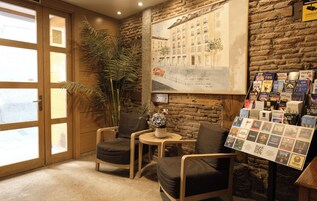 Una camera accogliente con pareti in legno, una grande finestra, un’area salotto con due poltrone, un tavolino con un vaso, e una libreria a muro con libri e brochure.