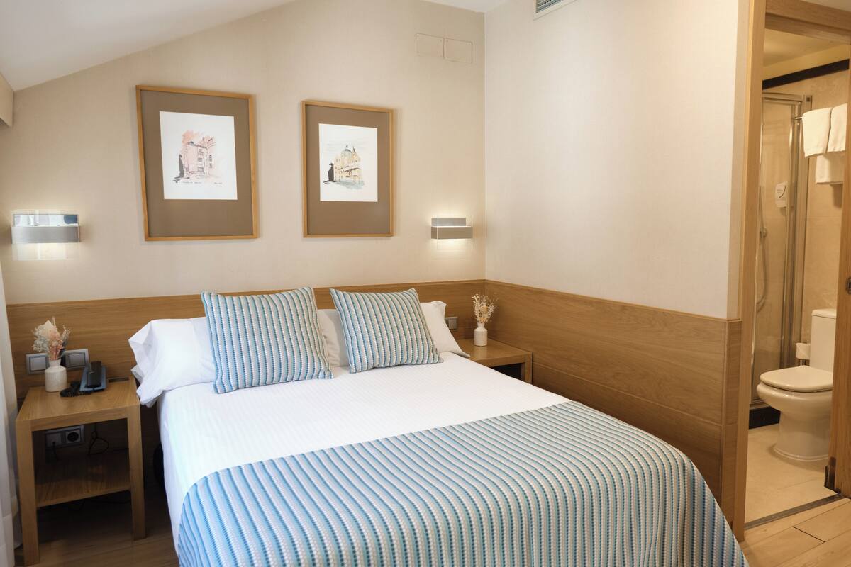 A hotel room with a large bed, two framed pictures on the wall, a nightstand with a lamp, and a wooden headboard.