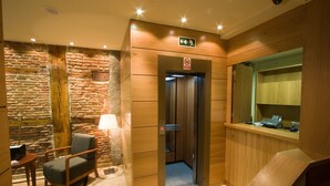 Un hall d’hôtel avec un mur en pierre, un comptoir d’accueil en bois, une chaise et un ascenseur.