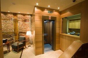 Una hall d'albergo con parete in pietra, una reception in legno, una sedia e un ascensore.