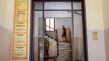 Ein Hotel Eingang mit einer Treppe, die zum zweiten Stock führt. Schilder weisen auf Hotel Berna und Silver Feathers hin, die Zimmerverfügbarkeit signalisieren.