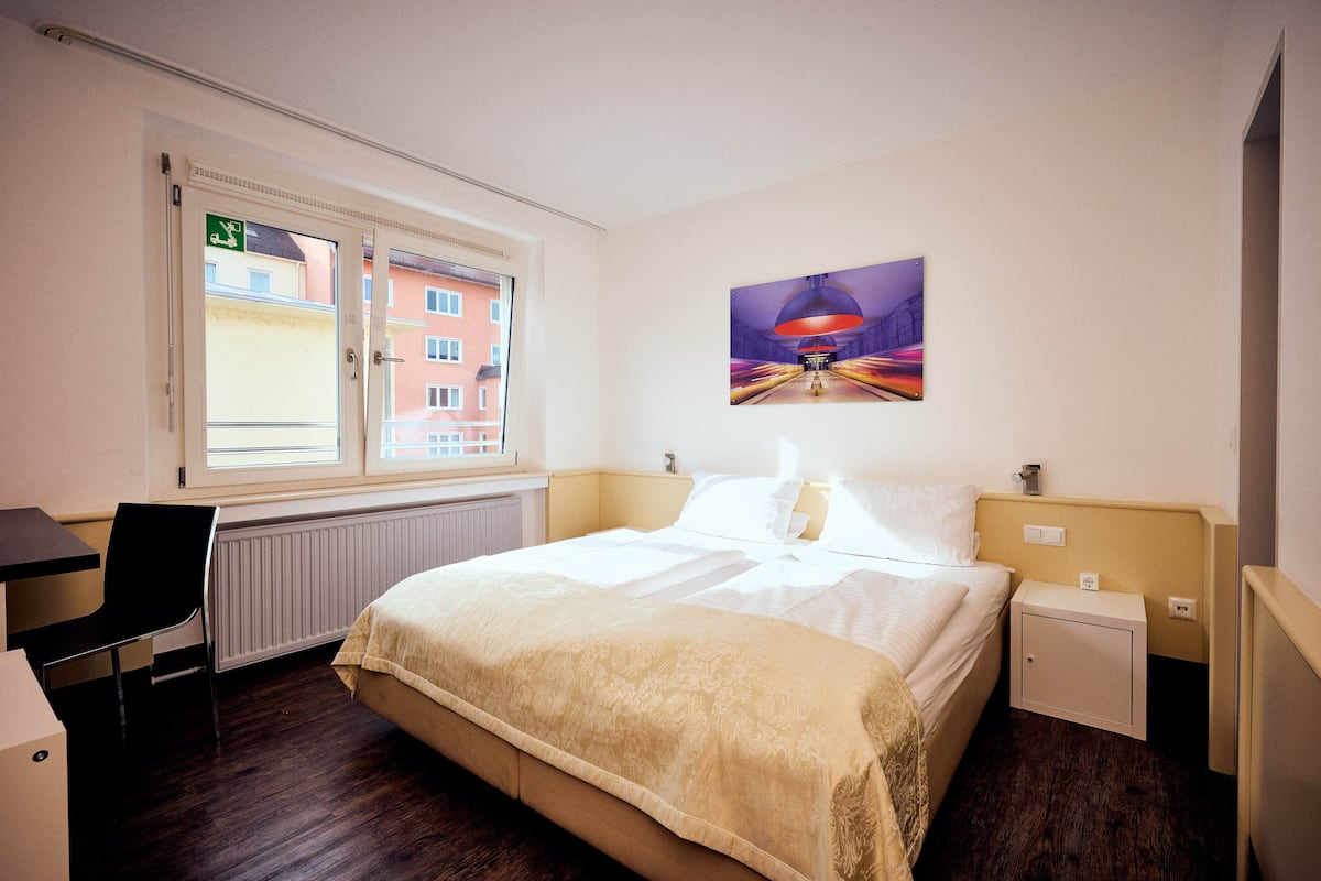 Ein Hotelzimmer mit Bett, Schreibtisch, Stuhl, Heizkörper und einem Fenster mit einem Notausgangsschild.