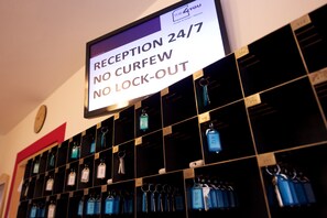 A reception area with a sign indicating reception is open 24/7, no curfew, and no lock-out. Key storage is organized in a grid with numbered tags. - The 4 You Hostel & Hotel (Munich)