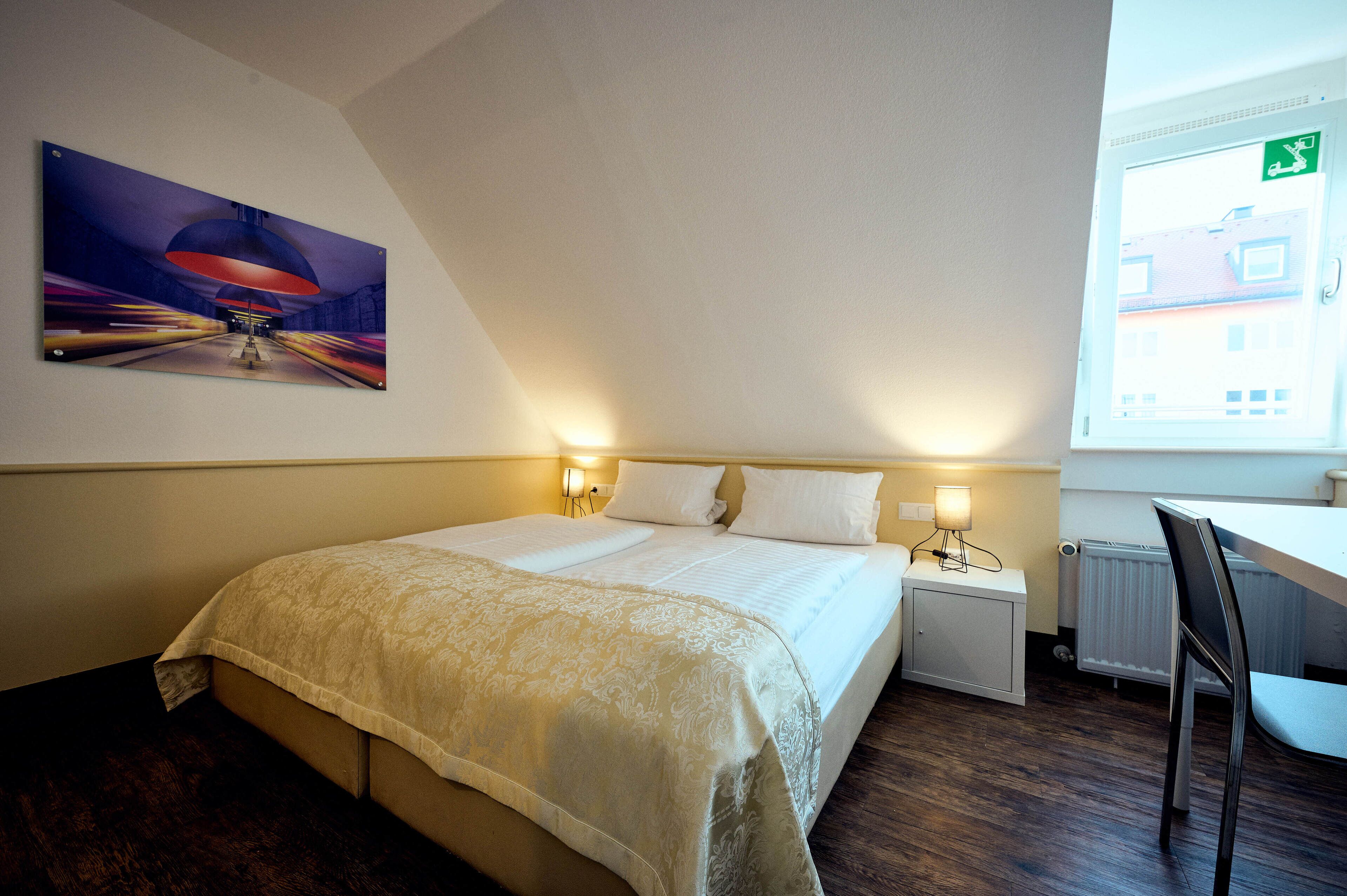 A hotel room with a bed, bedside lamps, a radiator, a desk, and a window with a view of buildings.
