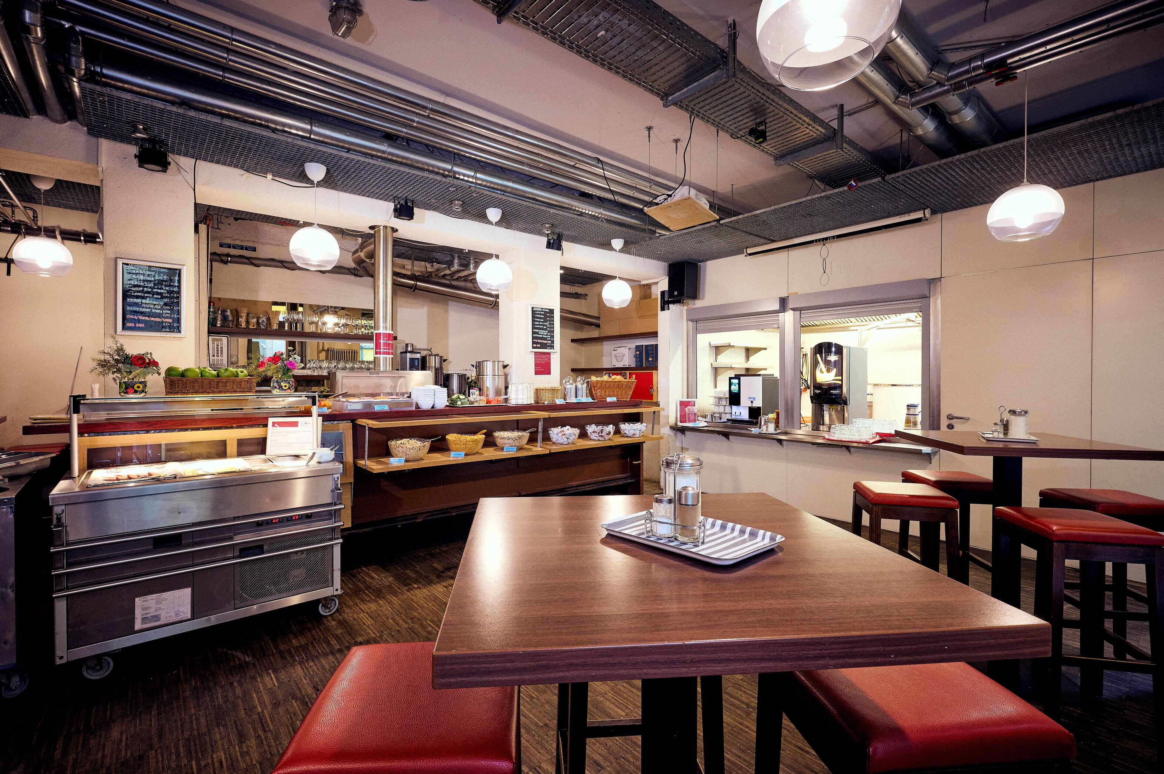 Ein Restaurantinneres mit einem Holztisch, roten gepolsterten Bänken, hängenden Pendelleuchten und einer Küche im Hintergrund.