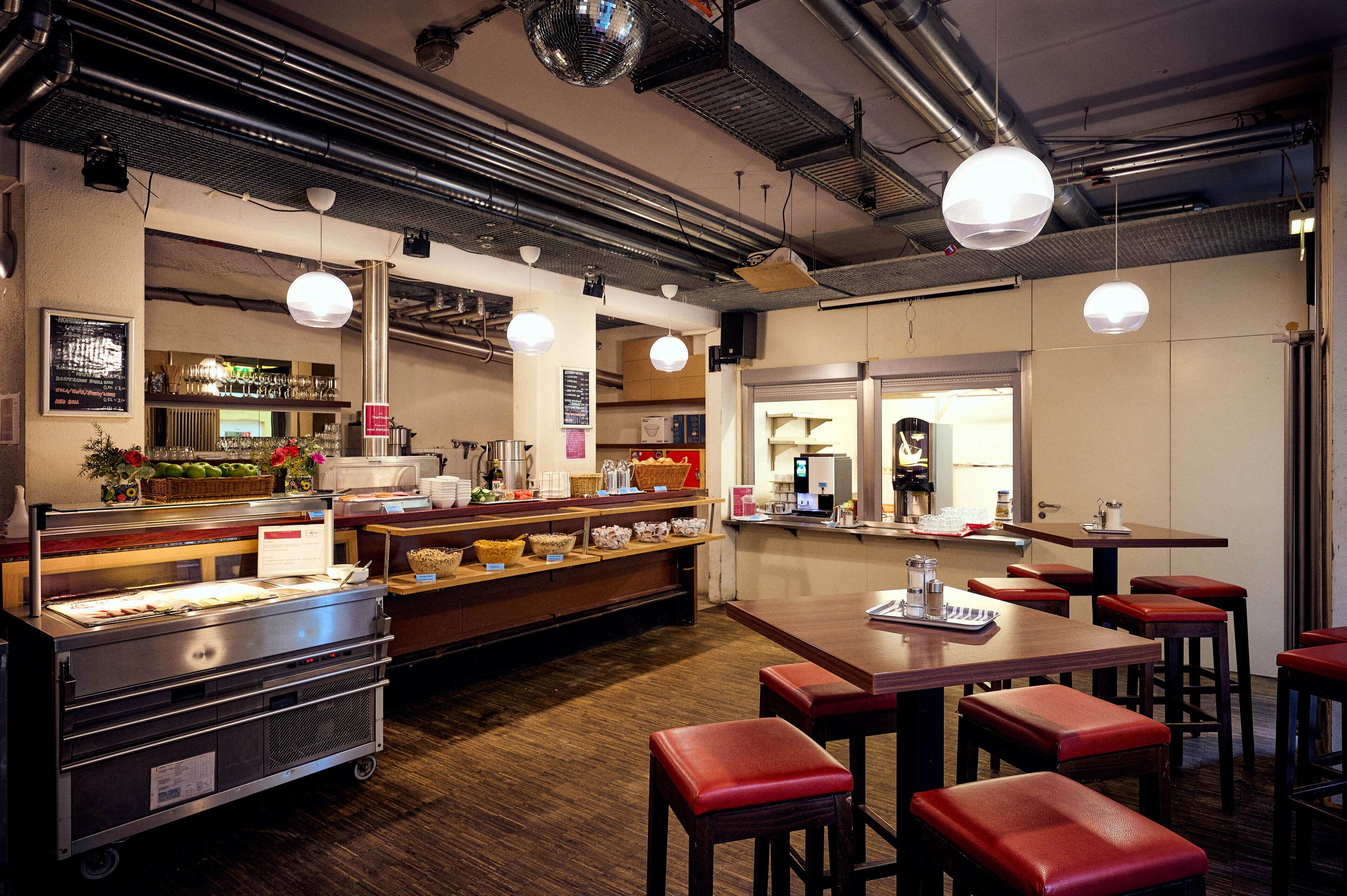 A restaurant interior with wooden tables, red cushioned stools, a buffet counter, and hanging pendant lights.