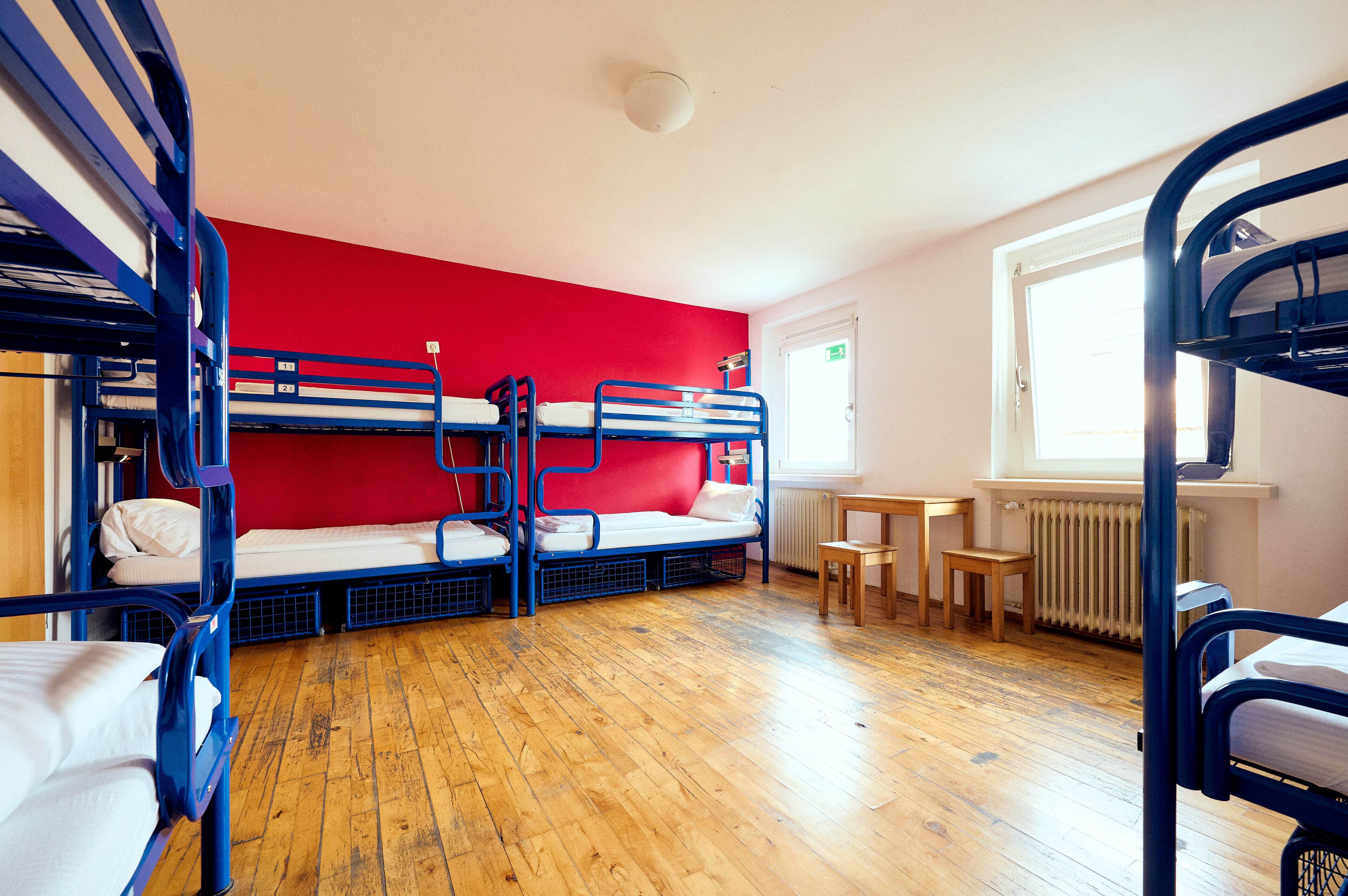 A dormitory room with bunk beds, a red wall, wooden flooring, a small table, and a window with a view.