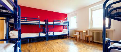 A dormitory room with bunk beds, a red wall, wooden flooring, a small table, and a window with a view.