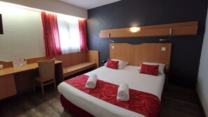 Ett hotellrum med en säng, ett skrivbord, en stol och ett fönster med gardiner.