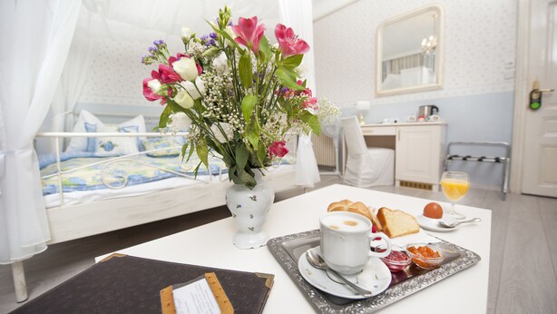 A hotel room with a bed, a bedside table with a tray of breakfast items, a vase of flowers, and a bathroom visible in the background. - Boutique Hotel Kugel Wien (Vienna)