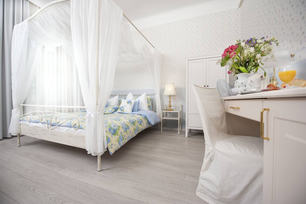 A bedroom with a four-poster bed, a bedside table with a lamp, a small chair, and a dresser with a mirror.
