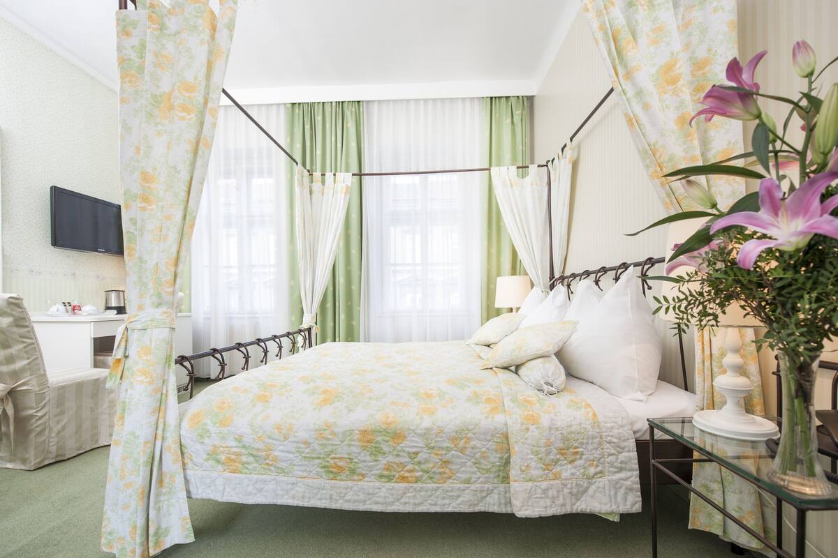 A bedroom with a four-poster bed, green curtains, a bedside table with a lamp, and a TV mounted on the wall.