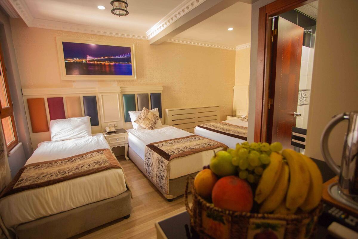 A hotel room with two beds, a nightstand, a lamp, a fruit basket, and a painting of a bridge.