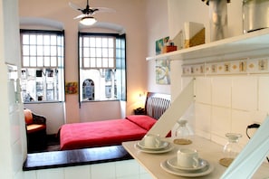 A compact hotel room with a bed, a small seating area, a dining table with tea set, and a view of the city through large windows.
