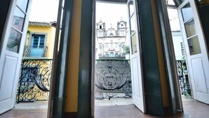 Un umbral abierto que da a un balcón con una reja de hierro forjado, un edificio amarillo e iglesia con dos torres al fondo.