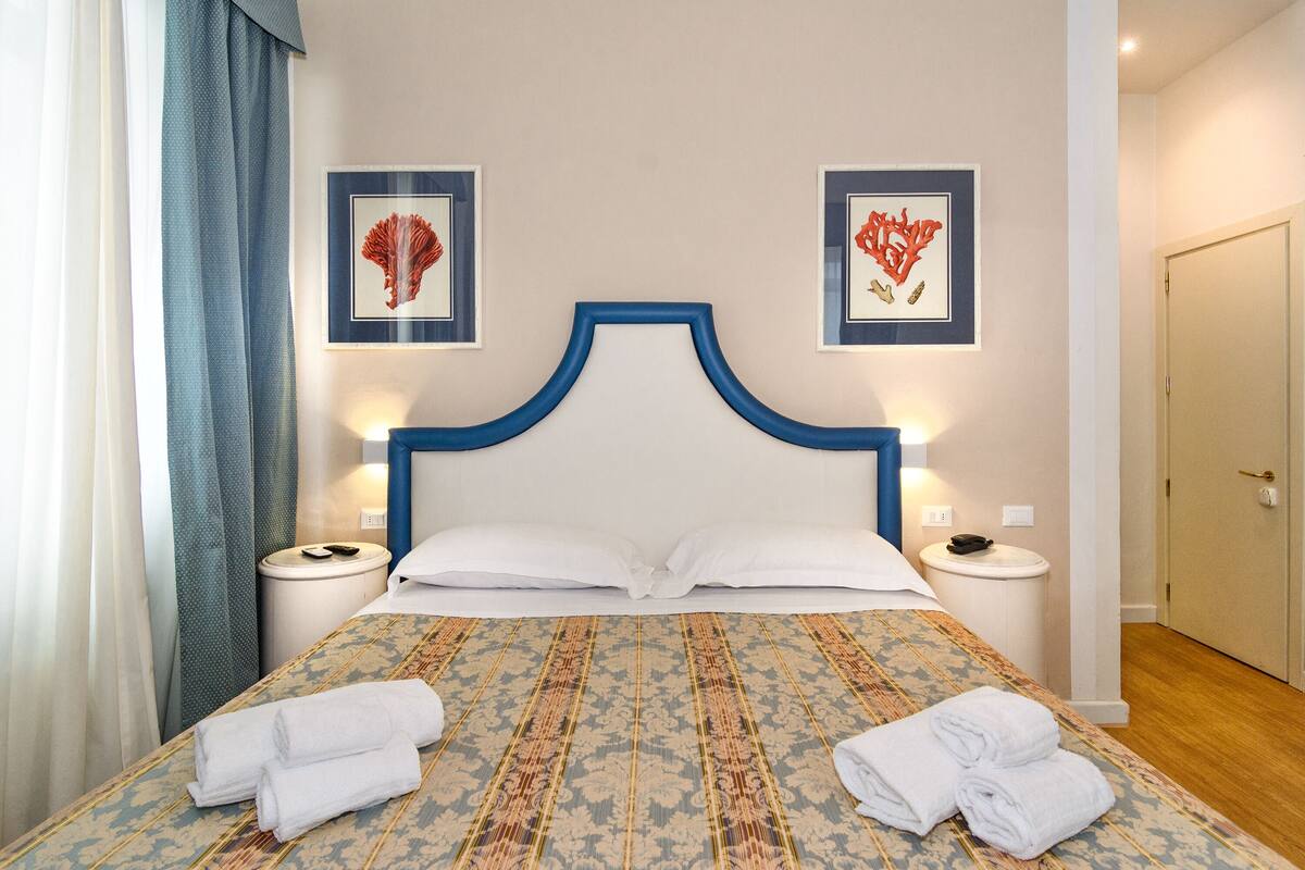Ein Hotelzimmer mit einem Bett, zwei gerahmten Kunstwerken, einem Nachttisch und einer Tür.