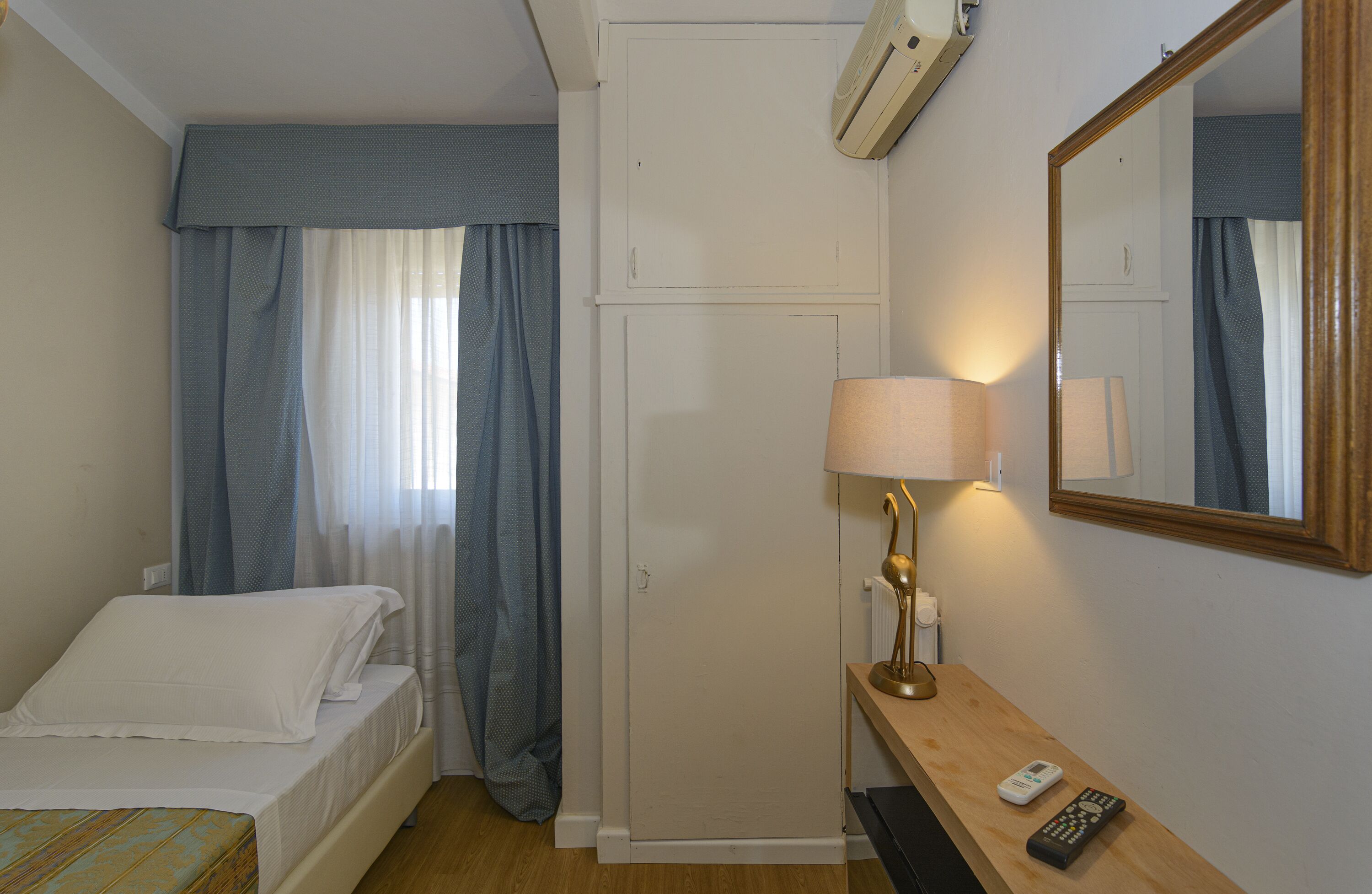 A hotel room with a bed, a desk with a lamp, a mirror, and a remote control.