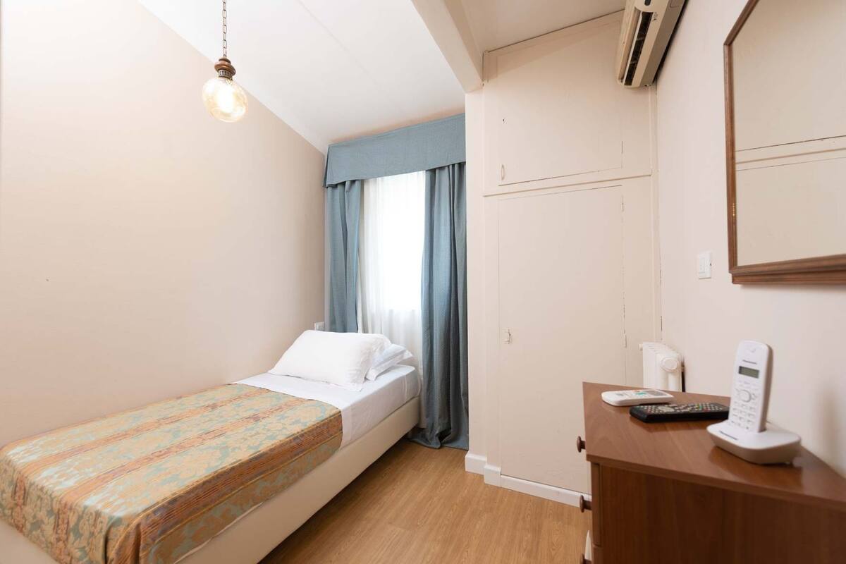 Ein kleines, ordentlich eingerichtetes Schlafzimmer mit einem Einzelbett, einem Nachttisch aus Holz, einem Telefon und einem Spiegel an der Wand.