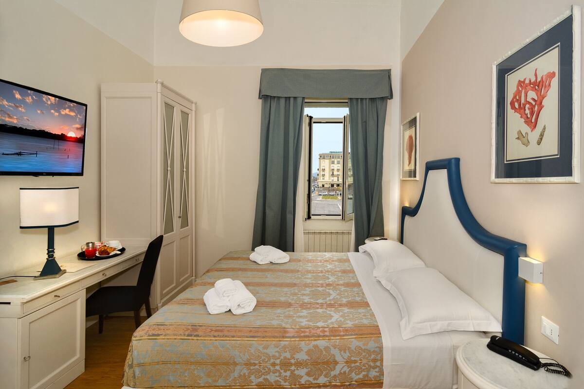 Ein Hotelzimmer mit Bett, Schreibtisch mit Lampe, Fernseher und einem Fenster mit Vorhängen.