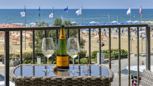 A balcony with a table featuring a bottle and two glasses, overlooking a beach with flags and umbrellas. - Hotel La Pace (Viareggio)