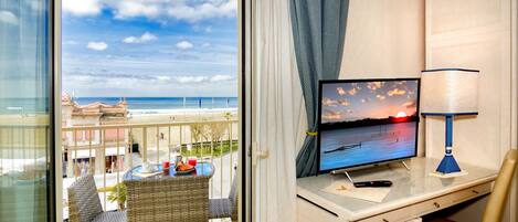A balcony with a view of the beach and ocean, a desk with a computer, a lamp, and a chair.
